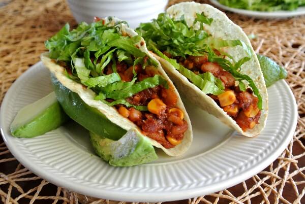 Slow Cooker Chipotle Tacos are two to a plate and leaning to the left. Filled with a veggies filling and topped with shredded lettuce and avocados.