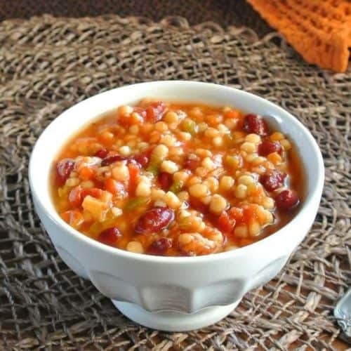 Slow Cooker Pearl Couscous Soup is in a white bowl with couscous, beans and carrots showing with the background being in dark brown.