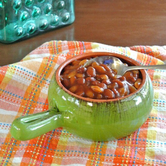 Crockpot baked beans are in a gren handled bowl on an old fashioned napkin.