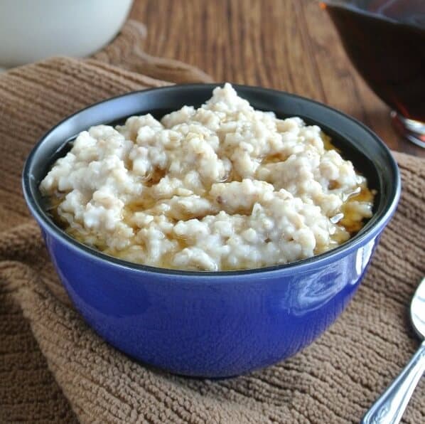 Slow Cooker Irish Oatmeal is filling a blue porcelain bowl with maple syrup floating on top. Spoon on the side.