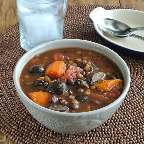 A bowlfull of rich dark soup filled with vegetables and legumes.