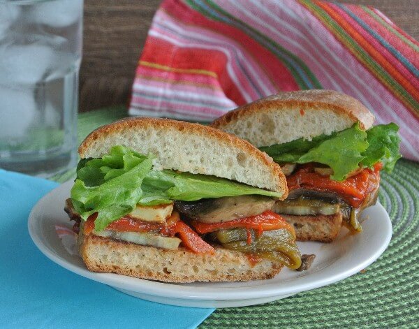 Stack of roasted vegetables on a wide bun and cut open to show the layers better and sitting on a white plate and turquoise napkin.