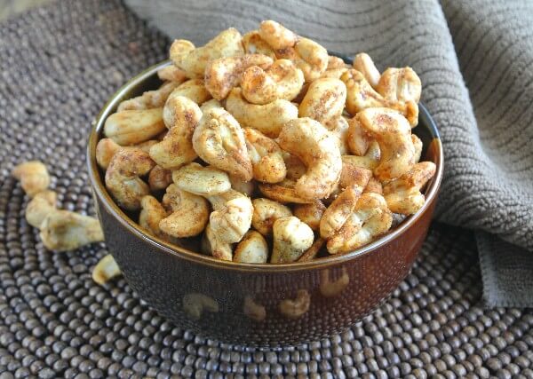 The Best Slow Cooker Spiced Cashews are golden and tilted forward in a chocolate colored bowl and is sitting on a dark brown beaded mat.