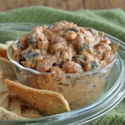 A side view of a clear bowl full of a rich brown chili dip with pita chips on the side.
