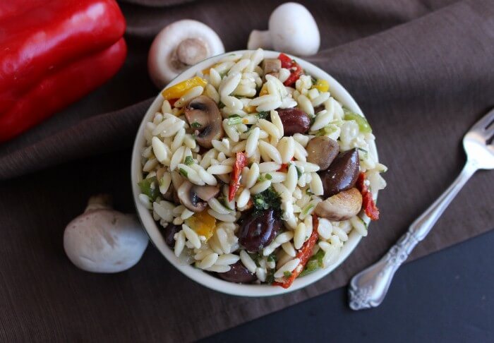 Overhead photo of lemon pasta salad showing olives and veggies. A fresh red bell pepper and mushrooms are scattered around the bowl along with a fork.