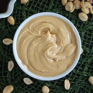 Overhead view of a white bowl holding creamy peanut butter with peanuts sprinkled around.