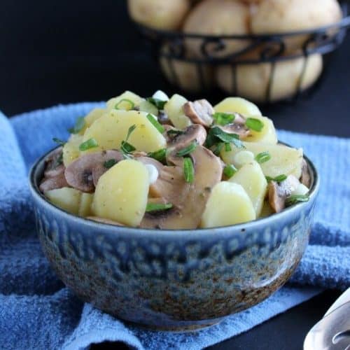 Pared potatoes and sliced mushrooms fill a blue speckled bowl with an iron basket of unpeeled potatoes behind.