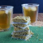 Almond Butter Protein Bars are sitting on a green mat and are cut into squares. Stacked 5 high infront of 2 glasses of iced tea.