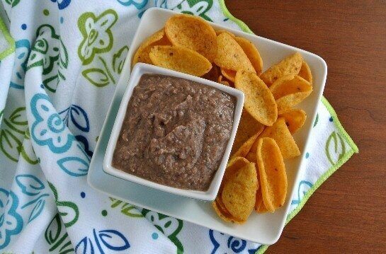 A square bowl full of dip is sitting on a square plate full of chips.