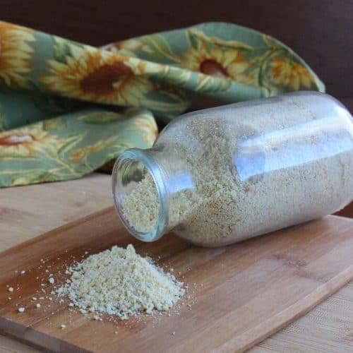 Vegan Parmesan Cheese is in a small glass jar and is laying on a wood cutting board. A little pile is in front of the glass so you can see the texture.