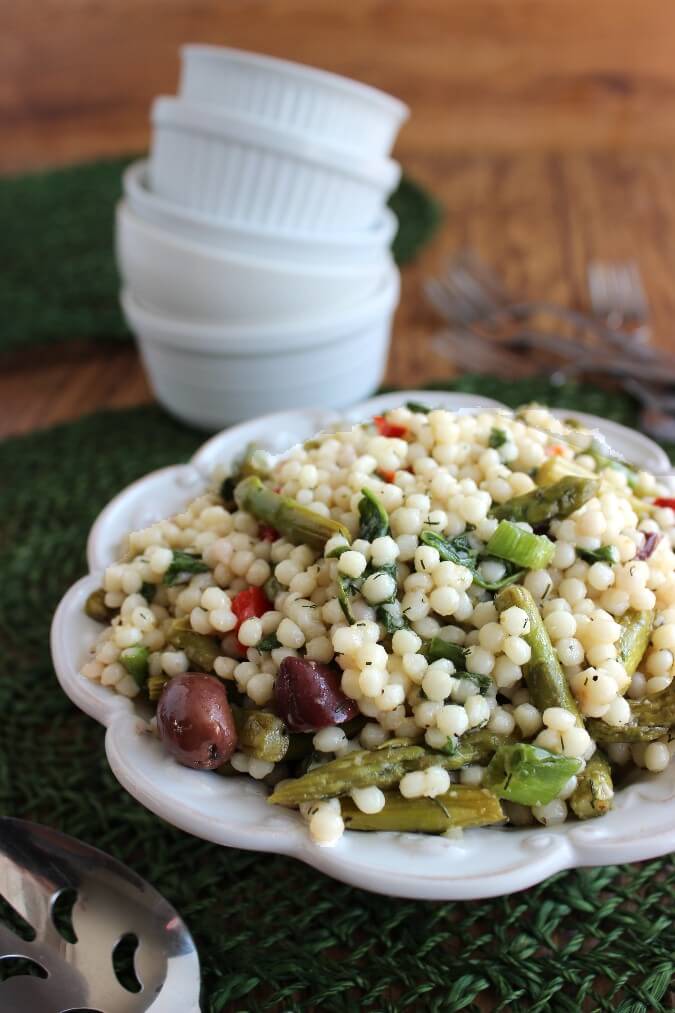 Cold salad is filling up a white scalloped bowl and the couscous has green asparagus and roasted red bell peppers peaking through this Pearl Couscous salad.