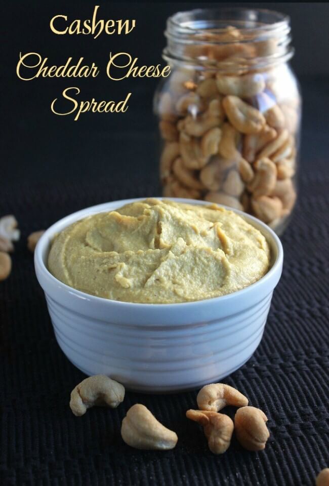 Cashew Cheddar Cheese Spread is a lovey pure golden shade and is piled in a white bowl on top of a black mat. Cashews are sprinkled on the mat and in a glas jar behind.