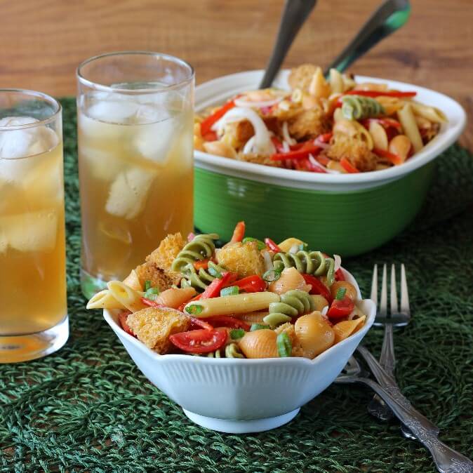 A white salad bowl is filled with colorful pasta, veggies and croutons. All against a green mat and a glass of iced tea.
