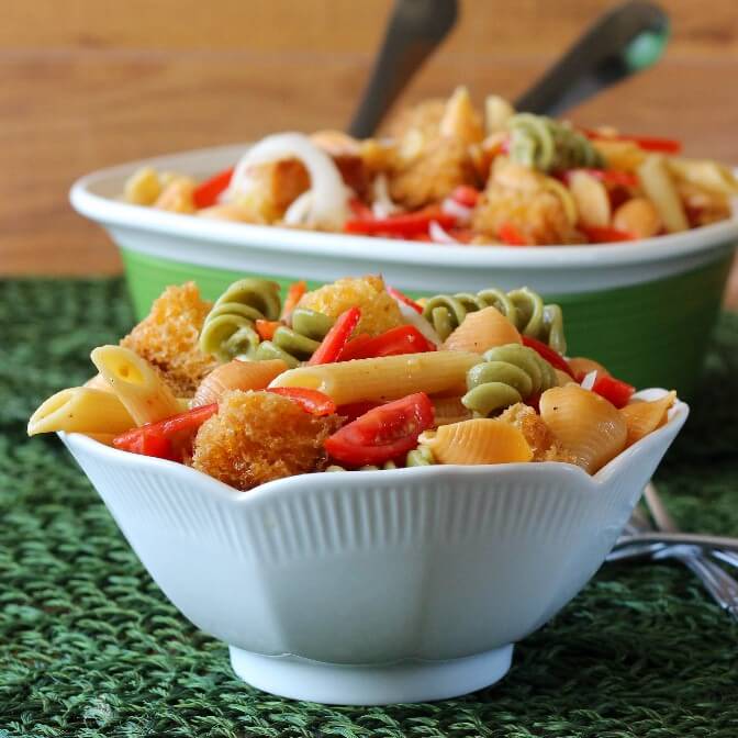 A white salad bowl is filled with Panzanella Pasta Salad in bright colors. A green and white serving bowl is filled with more in the rear.