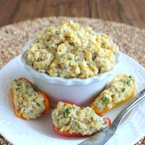 Three mini pepper halves are stuffed with a chickpea salad filling and sitting on a white plate in front of a bowlful of the filling.