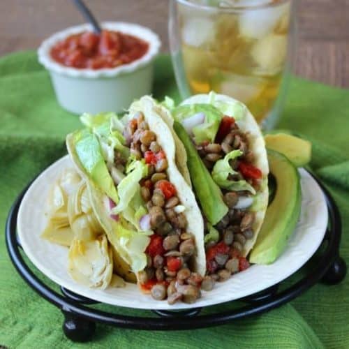 Two spicy lentil tacos are leaning against each other on a white [late with avocado and salsa on the side.