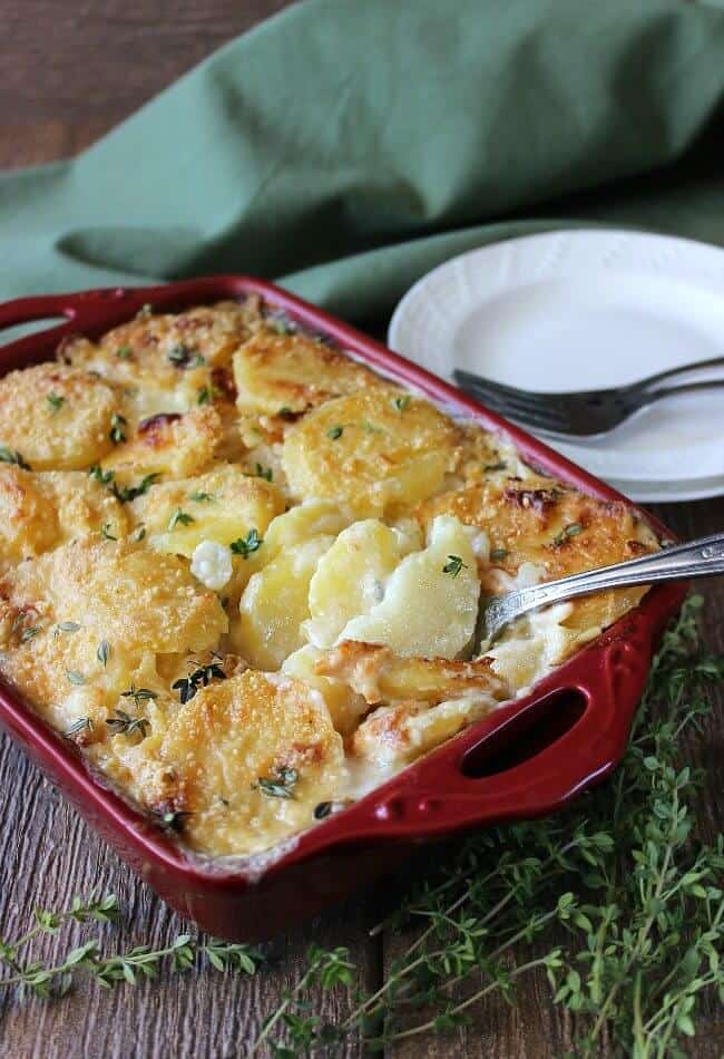 Dairy Free Herbed Scalloped Potatoes cotte a un colore dorato e servite in una casseruola rossa. Un grande cucchiaio per servire la prima porzione.