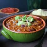 Two Bean Camp Chili is front and center in a green bowl siting on a brown cloth napkin. Rich red beans are topped with triangle cut avocado.