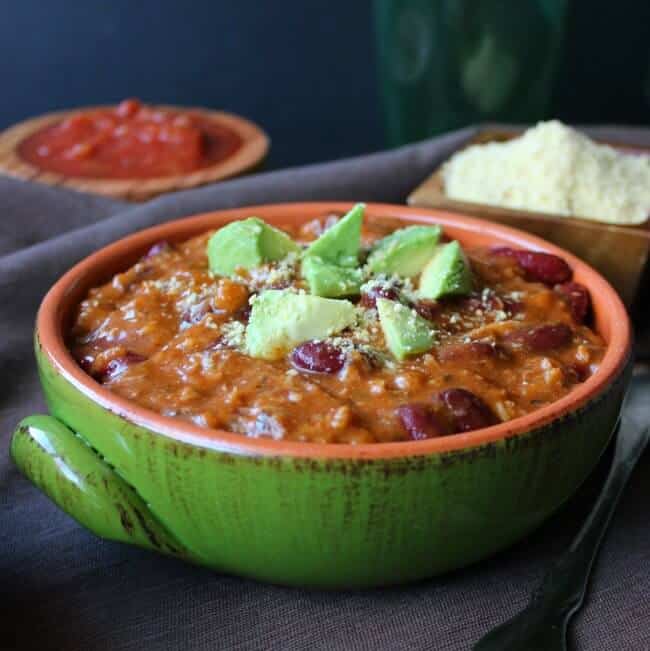 Two Bean Camp Chili is front and center in a green bowl siting on a brown cloth napkin. Rich red beans are topped with triangle cut avocado.