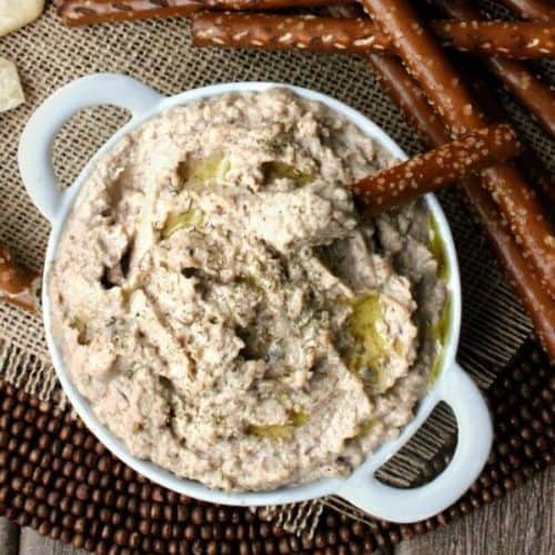 Kalamata hummus is in an overhead view in a small white handles bowl with a pretzel rod scooping up a bite.