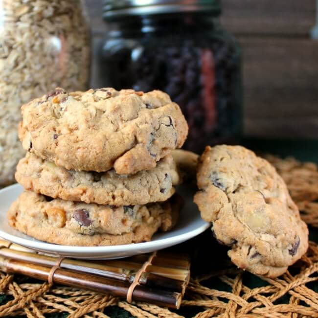 Nut Free Oatmeal Chocolate Chip Cookies are stacked three high on a small white plate and a woven mat. One cookie is set on the side at an angle.