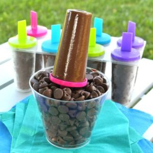 Chocolate ice cream on a stick is sitting upright in a clear glass of chocolate chipes with more fudgsicles in the background.