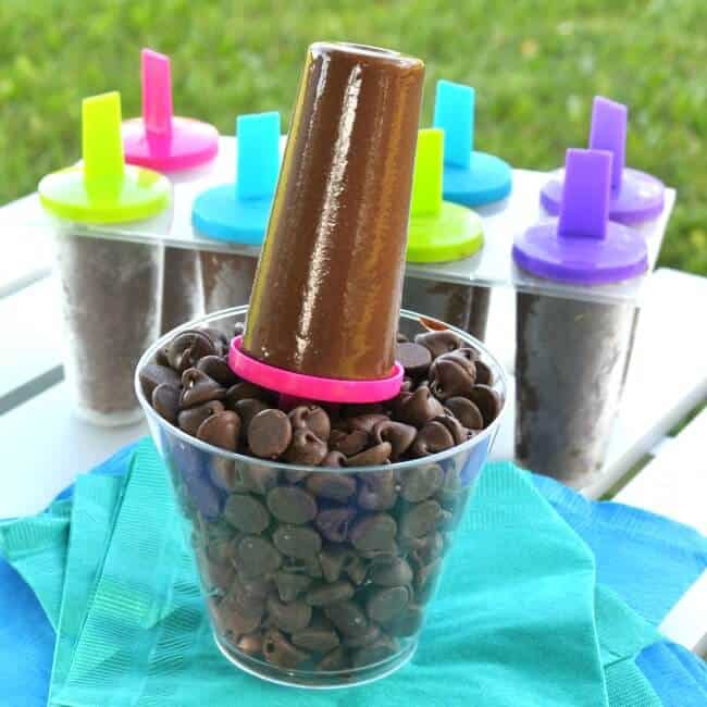 Chocolate ice cream on a stick is sitting upright in a clear glass of chocolate chipes with more fudgsicles in the background.