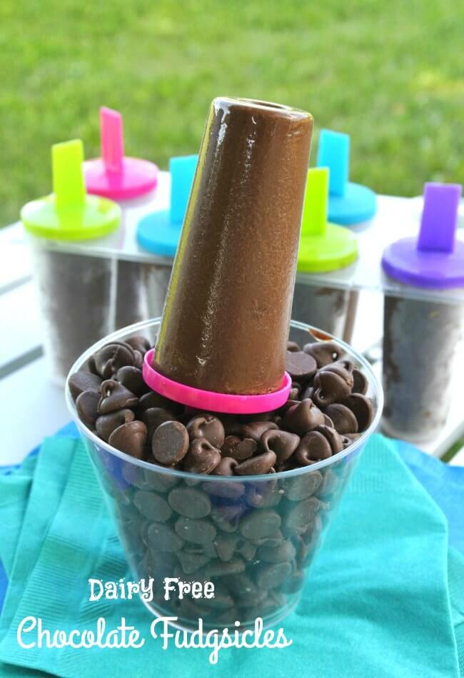 Chocolate ice cream on a stick is sitting upright in a clear glass of chocolate chips with more fudgsicles in the background.