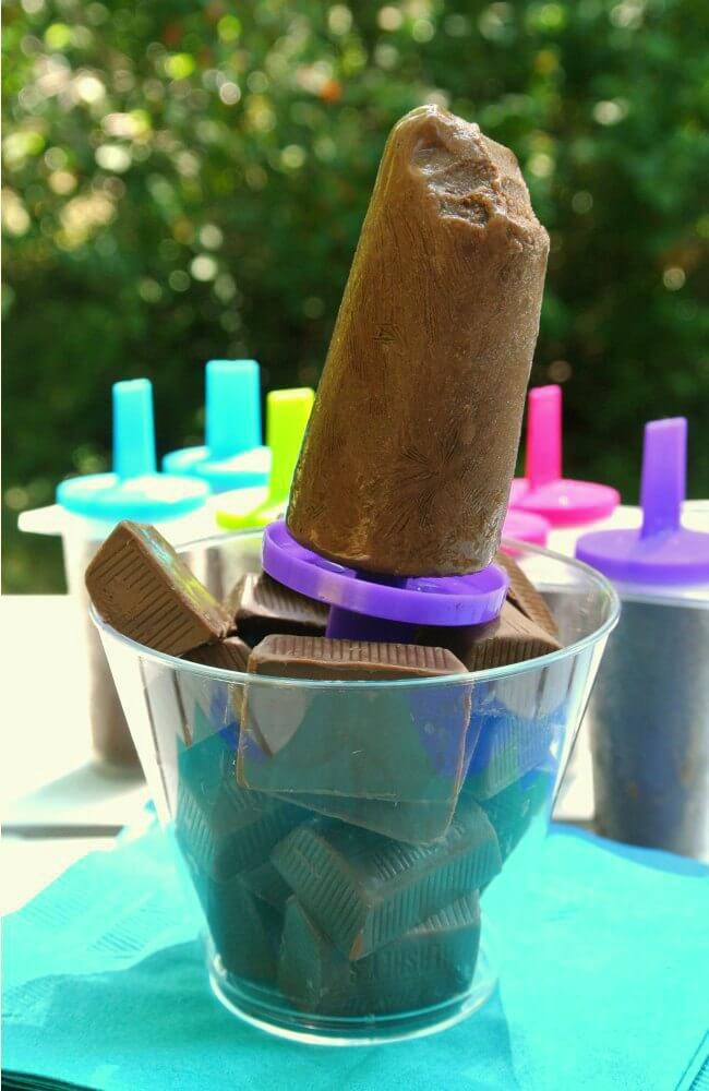 Chocolate ice cream on a stick is sitting upright in a clear glass of hershey squares with more fudgsicles in the background.