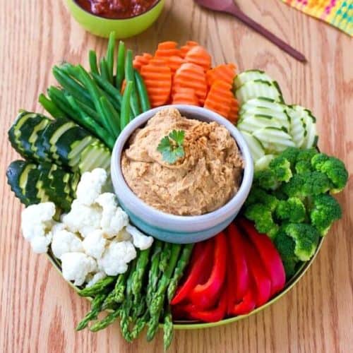 White bean dip is in a white bowl and is also surrounded of groups of sliced fresh veggies. All are on a round plate.