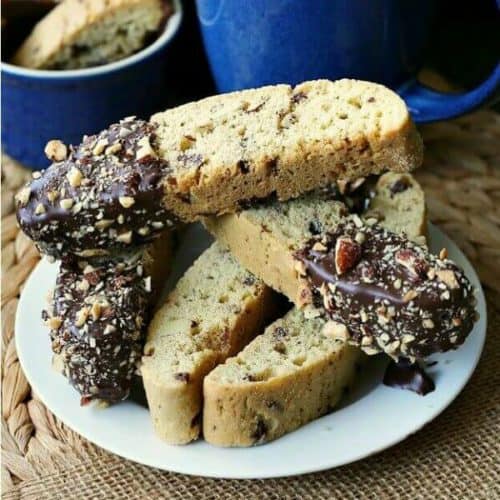 Chocolate Dipped Vanilla Biscotti are stacked diagonally and every which way on a white plate with chocolate dripping off the dunked end.