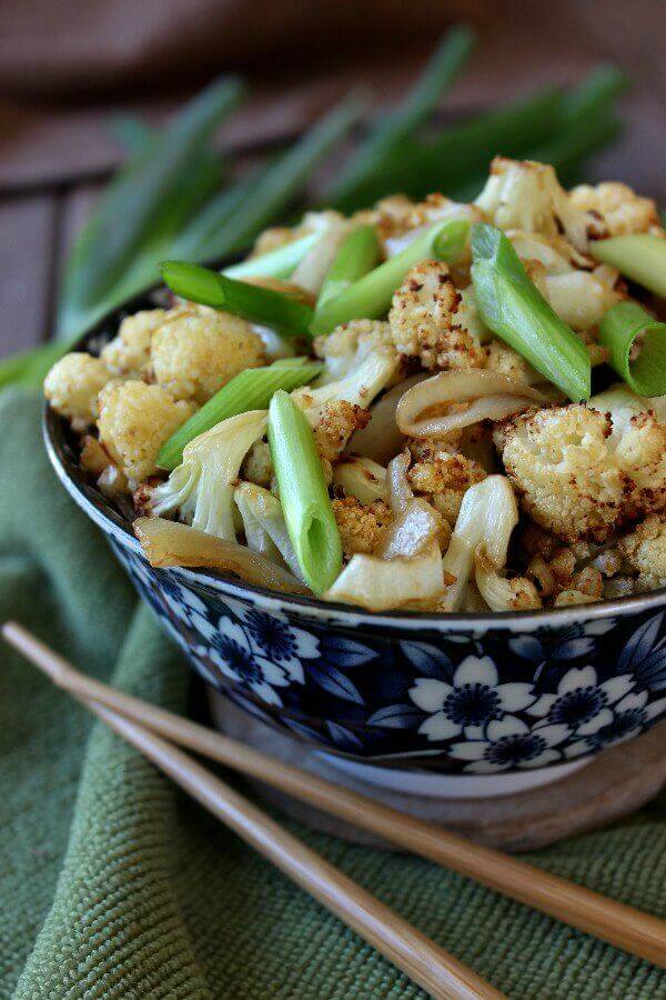 An Asian styled blue and white bowl cropped by thirds for the photo and the bowl is holding cooked cauliflower. It's garnished with scallions and chopsticks laying on the side.