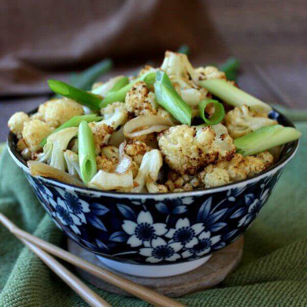 An Asian styled blue and white bowl holding cooked cauliflower garnished with scallions and chopsticks laying on the side.