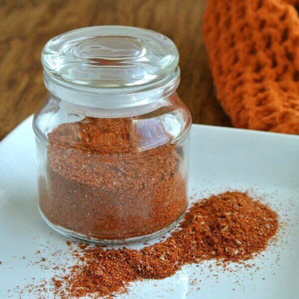 Homemade Taco Seasoning in rich rusty orange color that you can see trough a glass lidded jar. Spicy Recipe is also spilled on a white plate.
