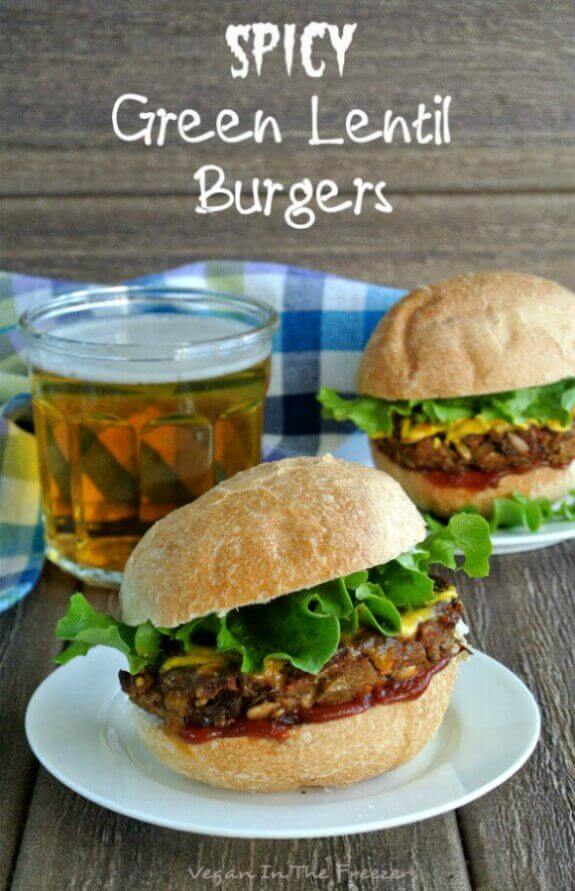 Fat lentil patty sitting on a bun with curly lettuce and condiments. Another sandwich is behind with a beer.