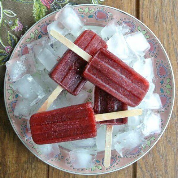 Blackberry and Grape Popsicles are stacked on top of each other on a bed of ice and a contrasting porcelain plate in purple, pinks and greens.