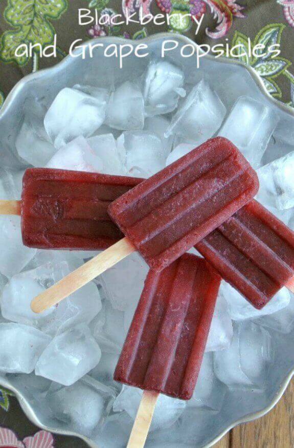 Blackberry and Grape Popsicles criss crossed and piled on a silver bowl filled with ice.