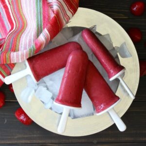 Four bright red frozen yogurt popsicles are on a bed of ice and a silver charger with a colorful striped cloth napkin on the side.