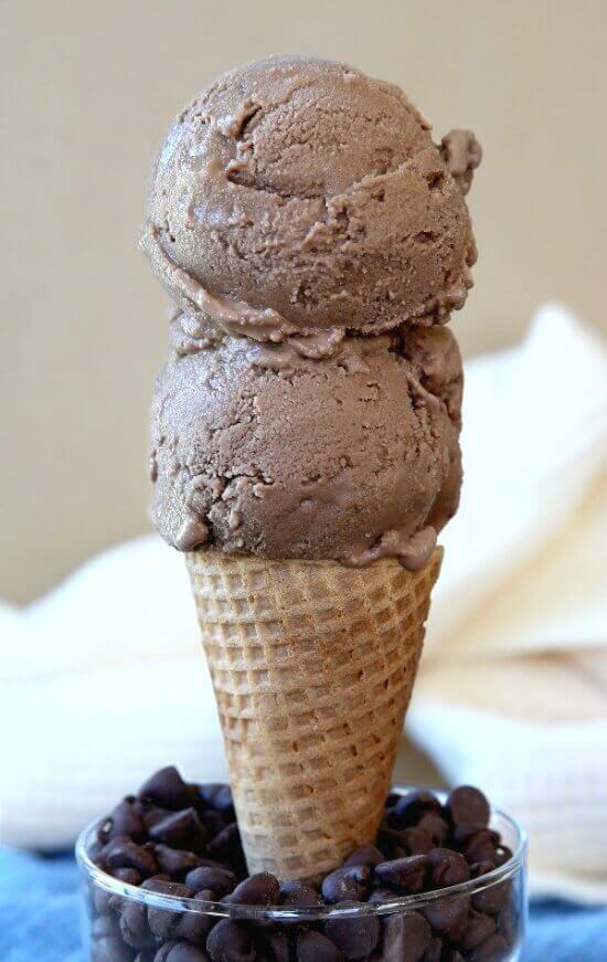 Two scoops of chocolate ice cream on a cone being held straight in a glass of chocolate chips.