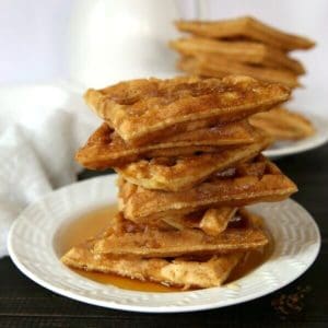Apple wafflesare stacked seven triangles high on a white plate with syrup.