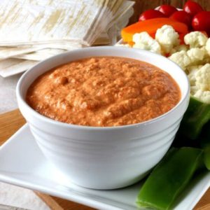 A thick red pepper dip is in a white bowl sitting on a white square plate with crudities around.