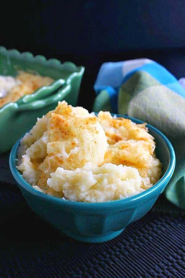 Mashed Potato Casserole has been spooned into a turquoise bowl with the casserole sitting behnd in a green pottery casserole.