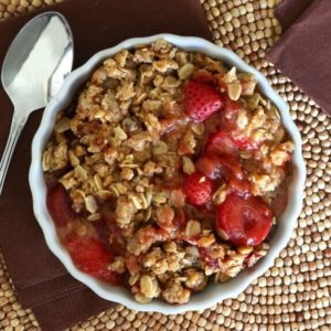 Slow Cooker Strawberry Crisp is center stage and fills a white scalloped bowl with red sliced strawberries and an oat and brown sugar topping.
