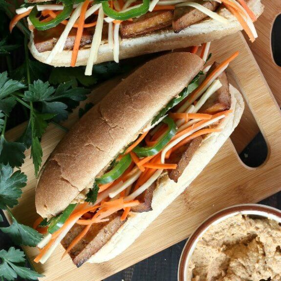 Two Long Sub Rolls laying on their back with layers of golden baked tofu with julienned carrots and parsnips and jalepenos. Bowl of spicy spread on the side.