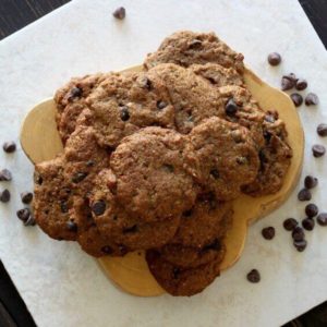 Chocolate Chip Banana Bread Cookies are stacked high on a pale wooden trivet with chocolate chips are scattered around.