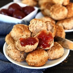 Cinnamon Cookie Biscuits are 2" across and are piled high on a white plate. Sitting on a navy cloth strawberry jam is waiting in a white square condiment bowl.