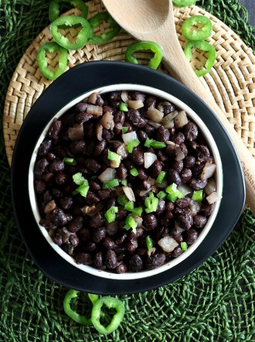  Slow Cooker Cuban Black Beans Recipe as an overhead view with black beans and onions piled in a white bowl and sprinkle with bright green jalapeño rings.