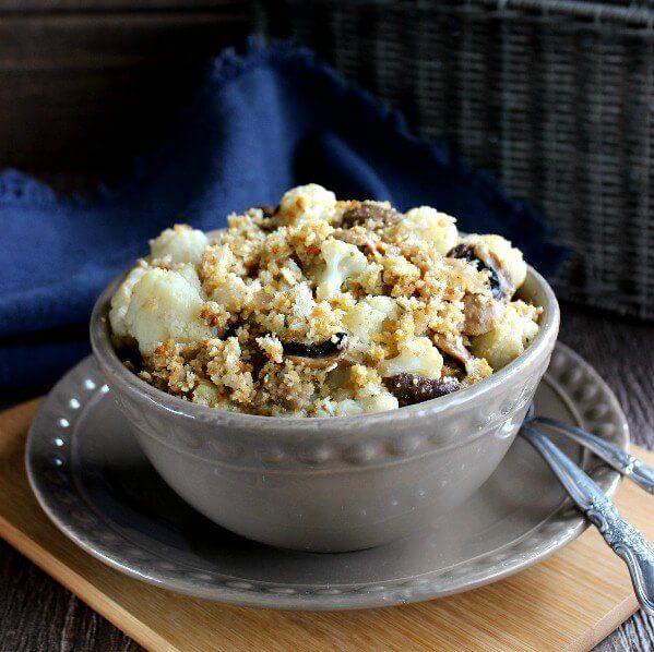 Cheesy Vegan Cauliflower Mushroom Casserole is piled high in a taupe bowl right in front of a navy cloth napkin. The bowl is tilted forward with delicious cauliflower and topping all mixed together.