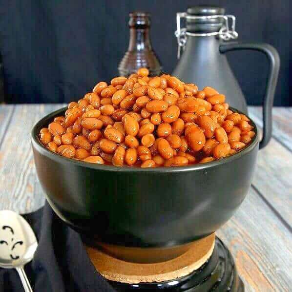 BBQ Baked Beans in their rust red color and piled high in a black bowl and sitting on a black trivet. Contrasting wood table in grays and rusty brown.