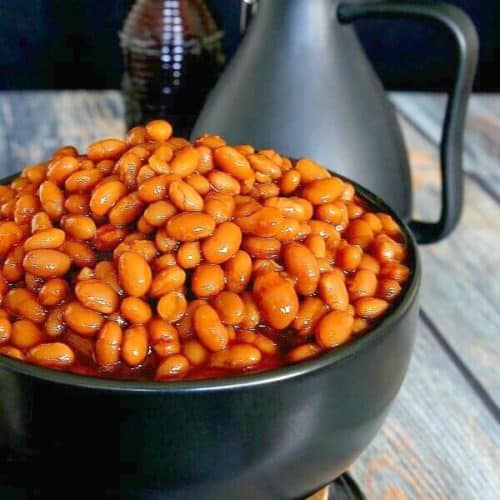 BBQ Baked Beans in their rust red color and piled high in a black bowl. Contrasting wood table in grays and rusty brown.